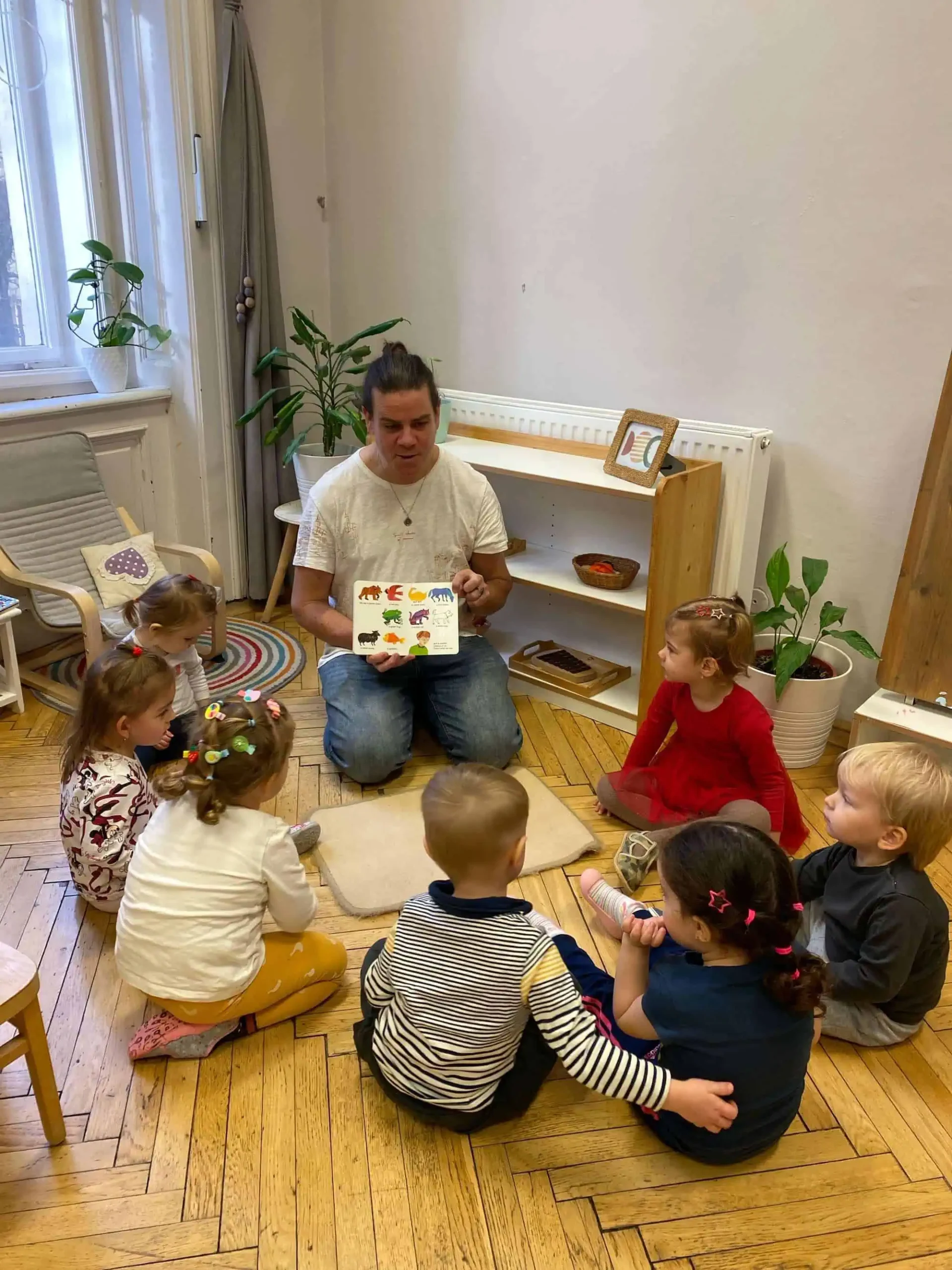 Učitelka sedí se zkříženýma nohama na podlaze a čte obrázkovou knihu skupině batolat sedících kolem něj v útulné třídě.